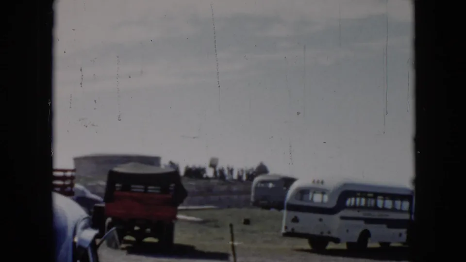 1956 Cuba 8mm frame with a white bus, red truck, older cars, stonework, and a distant group on a grassy elevated site.