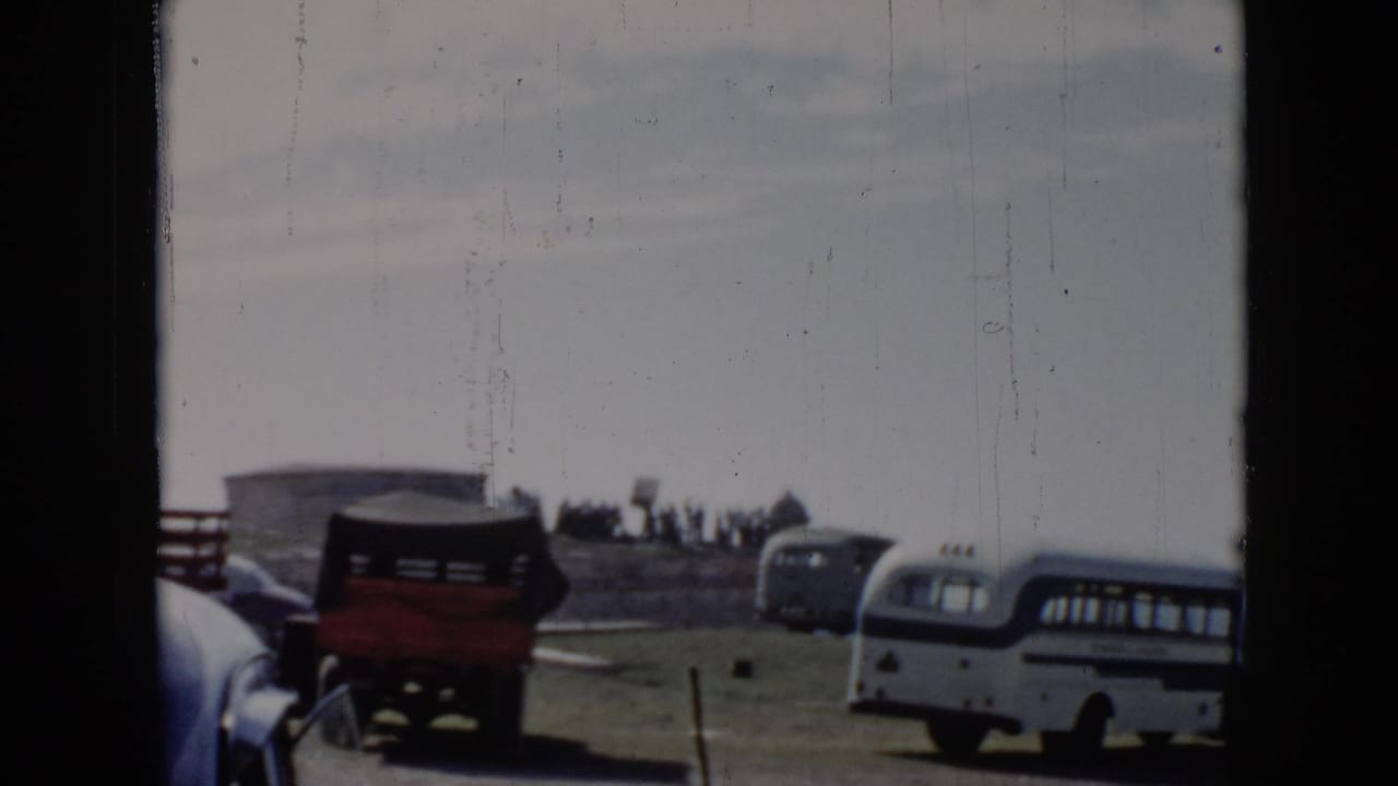 1956 Cuba home-movie frame showing a white and blue bus, red truck, older cars, and distant people near an ocean cliff-side area.