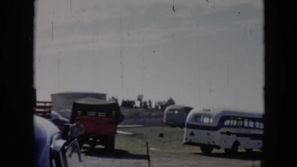 1956 Cuba frame with a red truck, white and blue bus, older cars, and small figures on a grassy cliff-side lot by the sea.