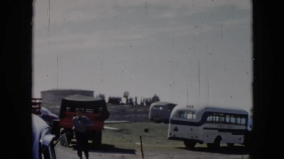 1956 Cuba home-movie frame showing a man running in front of a red truck, older cars, and a white and blue bus near the coast.