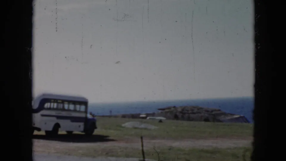 1956 Cuba coastal frame showing a white and blue bus on a dirt road, low cliff-side ground, and the ocean horizon in the distance.
