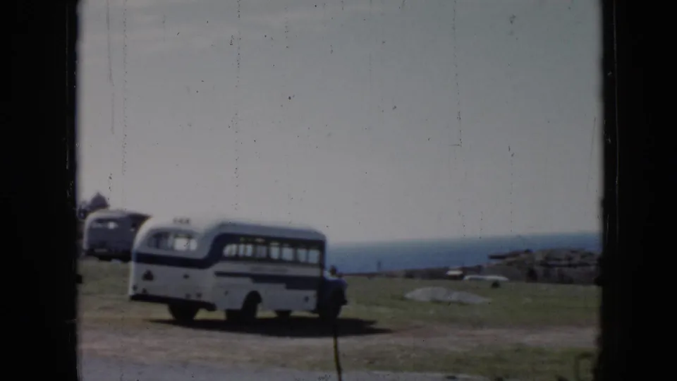 1956 Cuba home-movie frame showing a white and blue bus near a grassy cliffside overlook, filmed through dark frame edges.