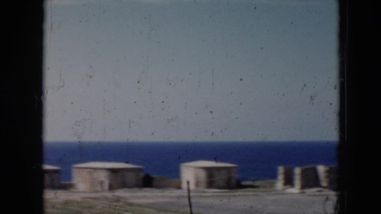 1956 Cuba 8mm frame showing a coastal scene through dark window bars, with low round structures near the shore and the sea behind them.