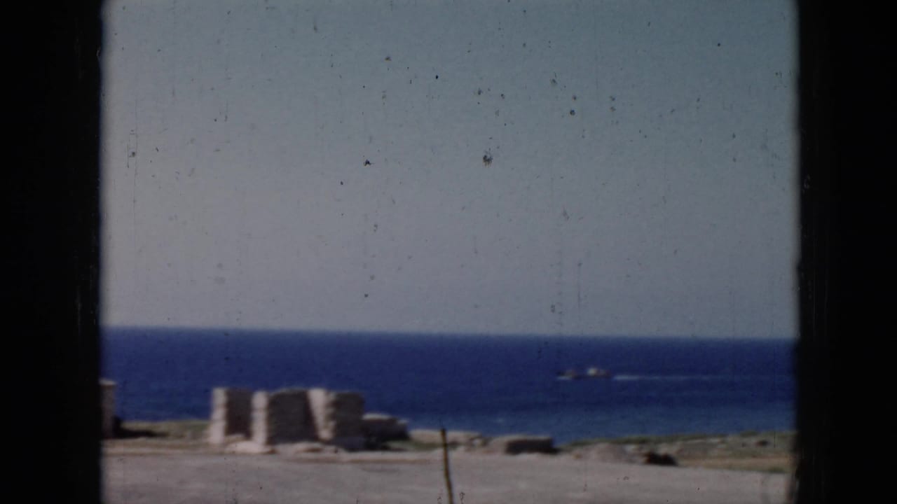 1956 Cuba home-movie frame looking through a window toward concrete shoreline structures, open water, and a small vessel on the horizon.