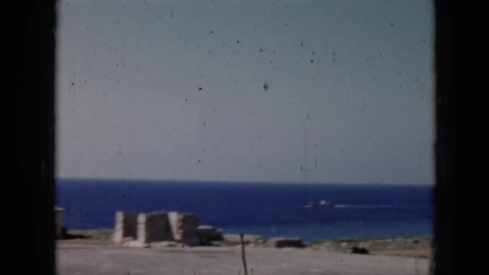 1956 Cuba home-movie frame looking through a window toward concrete shoreline structures, open water, and a small vessel on the horizon.
