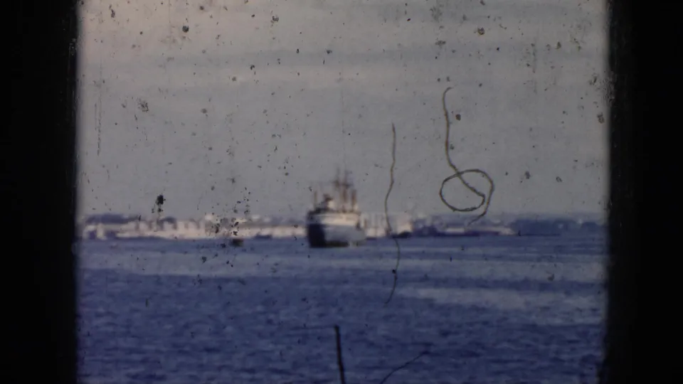 1956 Cuba home-movie frame with a ship offshore, dark window framing, blue sea, pale sky, and prominent scratches on the film surface.