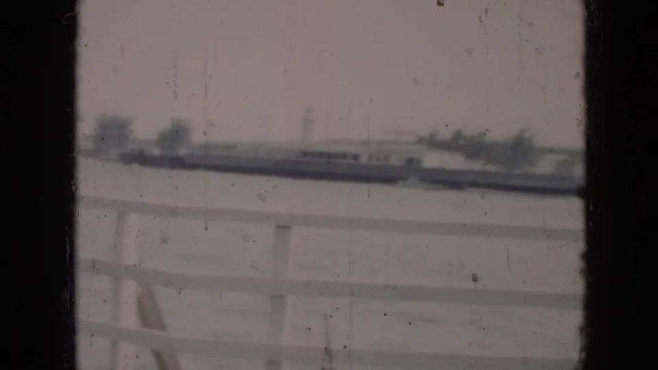 Mid-century Cuba home-movie frame looking past a white railing toward water, trees, and low buildings on the opposite shore.