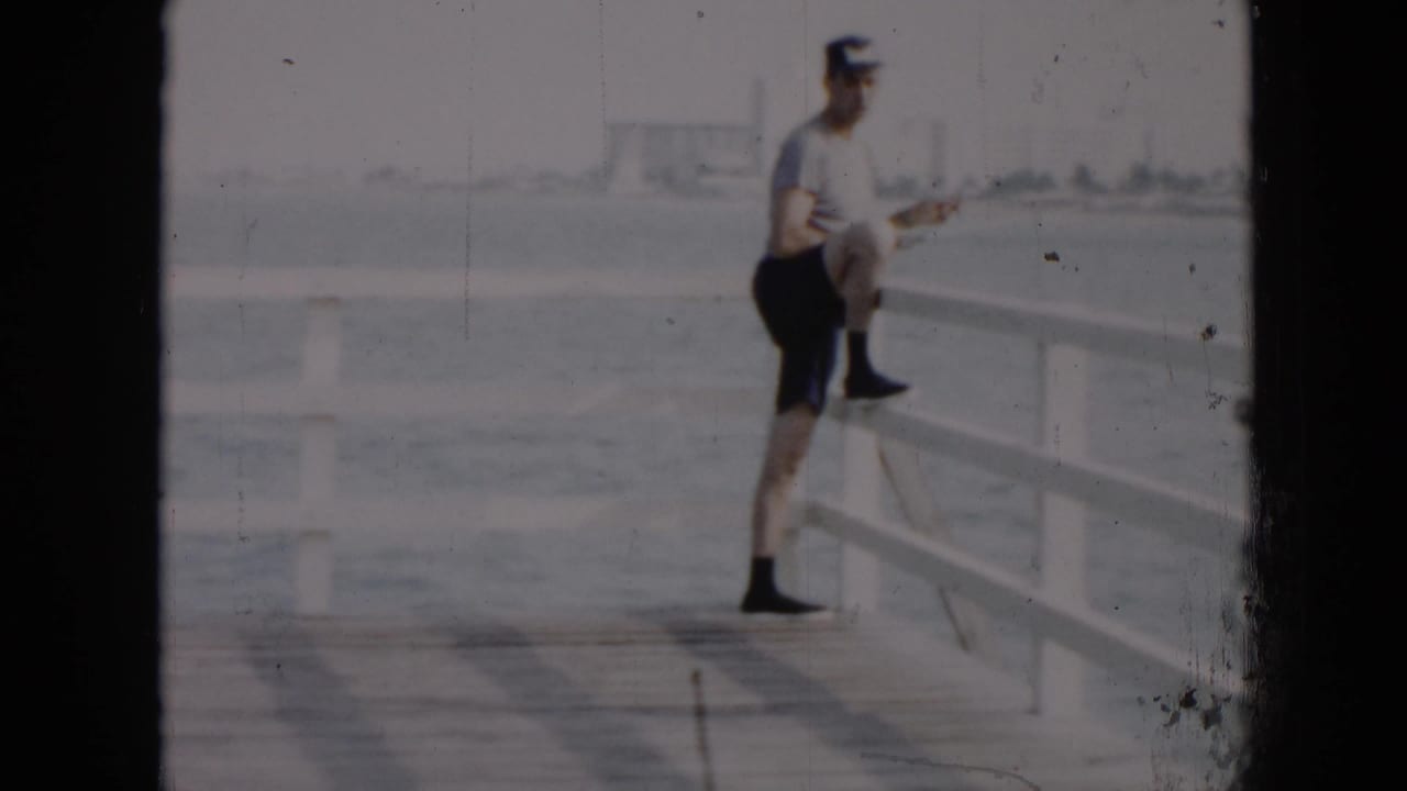 In Cuba in 1956, a man in a light shirt, cap, and dark shorts leans against a white pier railing by calm water.