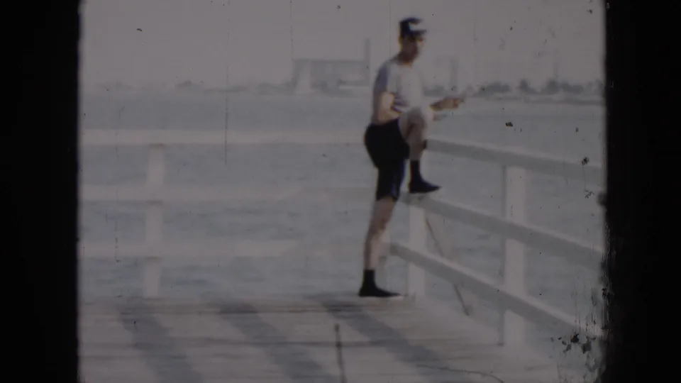 In Cuba in 1956, a man in a light shirt, cap, and dark shorts leans against a white pier railing by calm water.