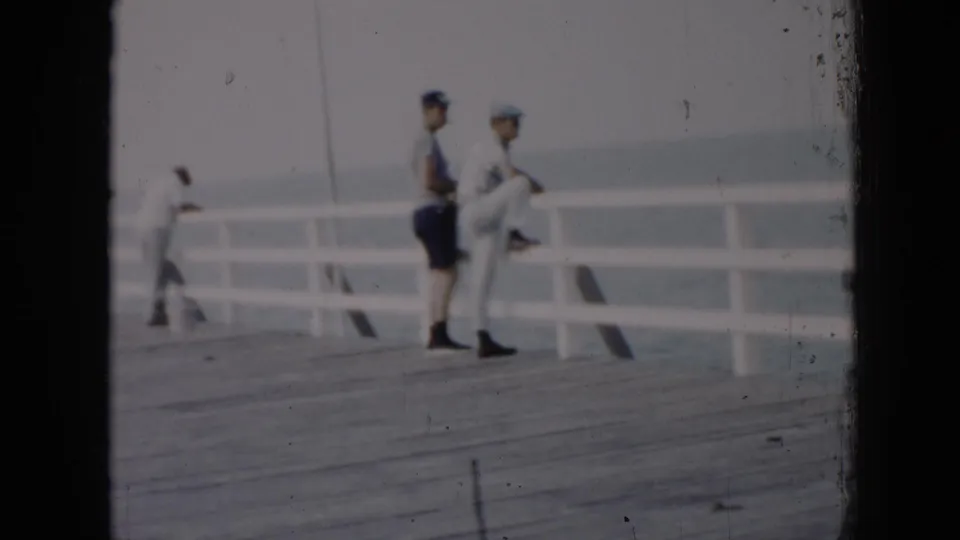 Two men in caps stand near a white wooden pier railing in 1956 Cuba, with calm water and another figure in the background.