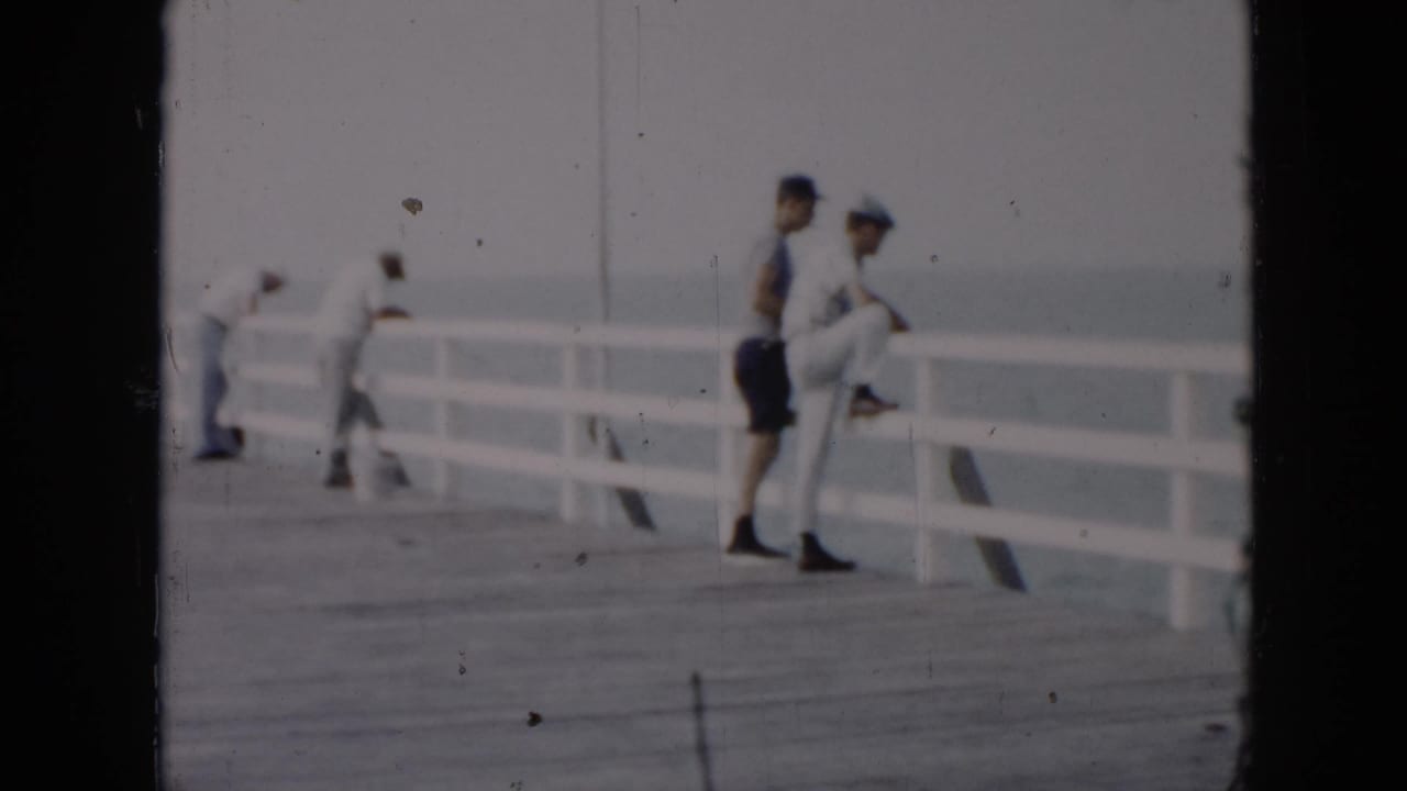1956 Cuba home-movie frame showing adults in summer clothing near a white pier railing beside calm water.