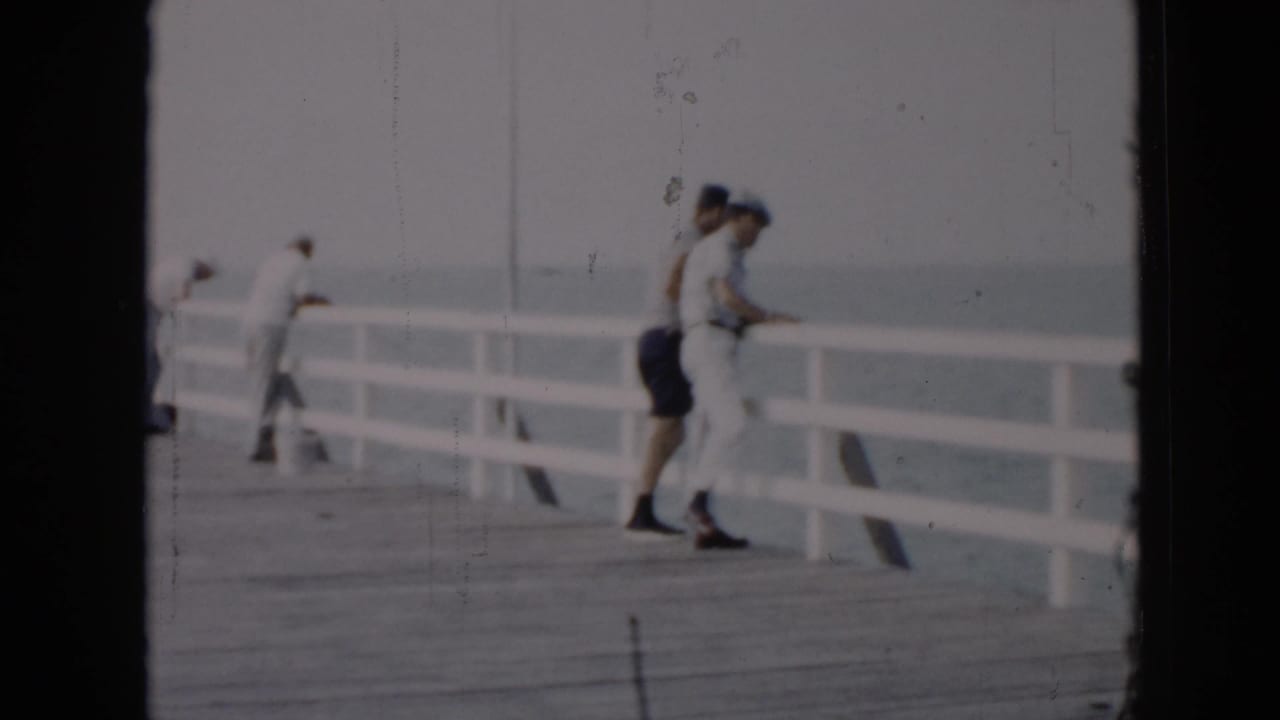 Two adults stand close to the white railing on a pier by the water.