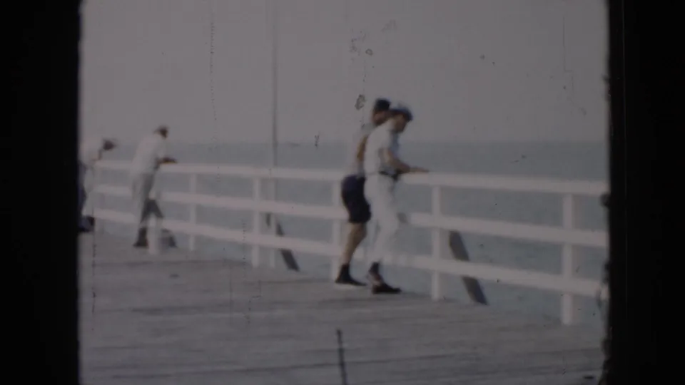 1956 Cuba frame with two adults near a white pier railing, one in light clothing and another in dark shorts, facing the ocean.