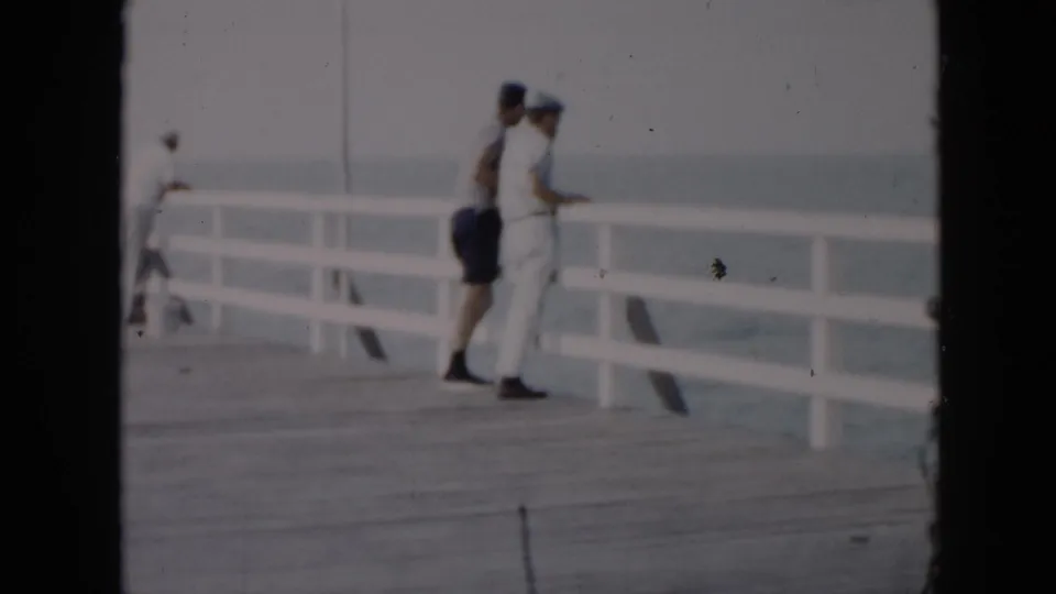 1956 Cuba home-movie frame showing adults along a white pier railing, with the ocean and hazy horizon behind them.