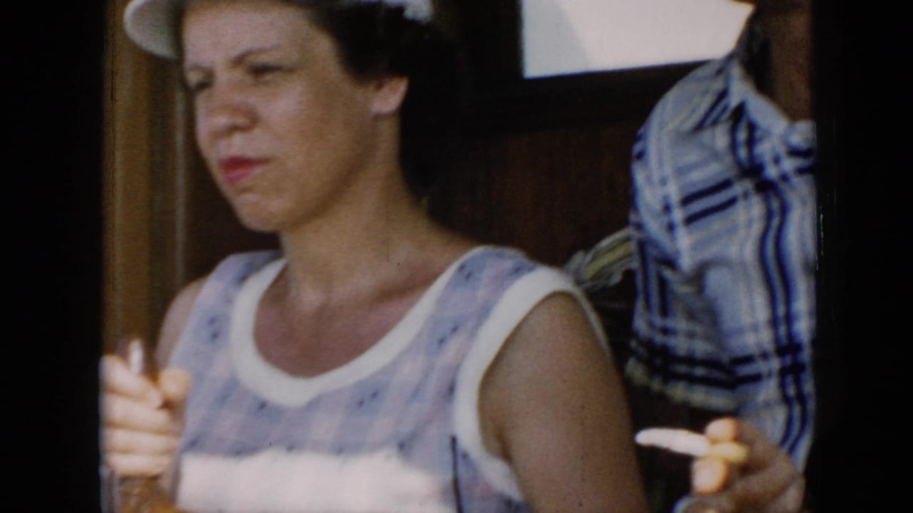 In 1956 Cuba home-movie footage, a woman in a sleeveless patterned dress stands near a wooden ship's wheel, partly framed by dark boat structure.