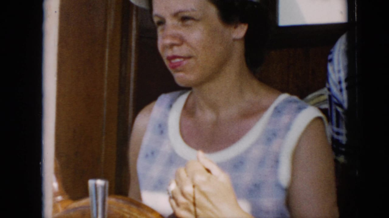 In supported 1956 Cuba footage, the woman stands by the wooden wheel, hands clasped near the controls, with boat woodwork behind her.