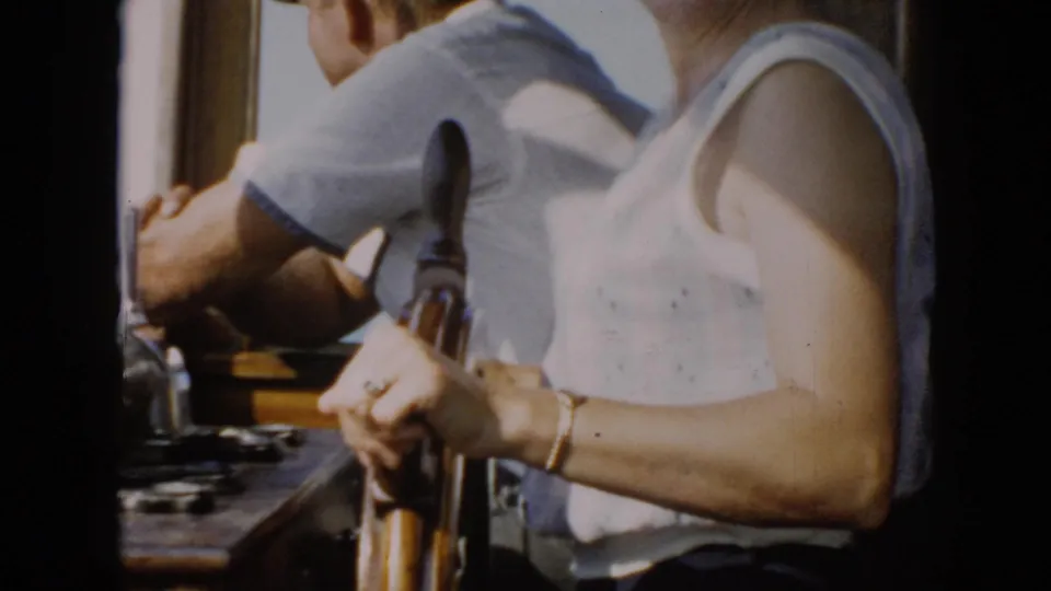 1956 Cuba 8mm frame showing a woman at a wooden ship wheel beside a man inside a sunlit bridge.