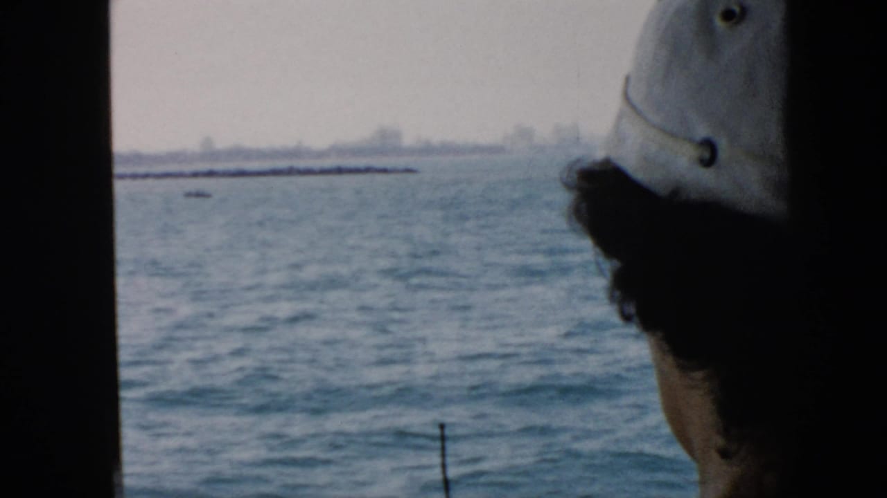 1956 Cuba home-movie frame showing a capped passenger in shadow looking out from a boat toward hazy water and distant land.