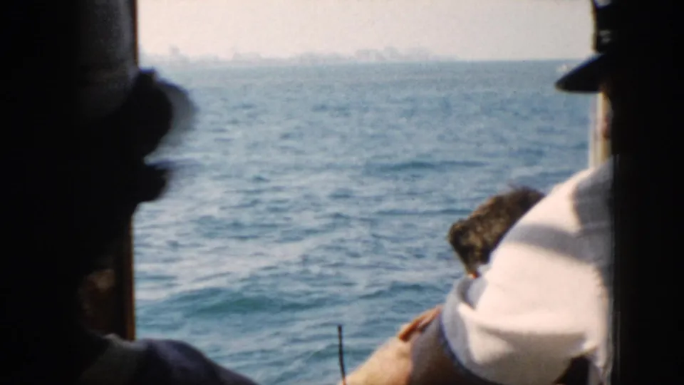 Boat passengers in shadow look toward the water, with a seated figure in a light shirt and hazy shoreline beyond.