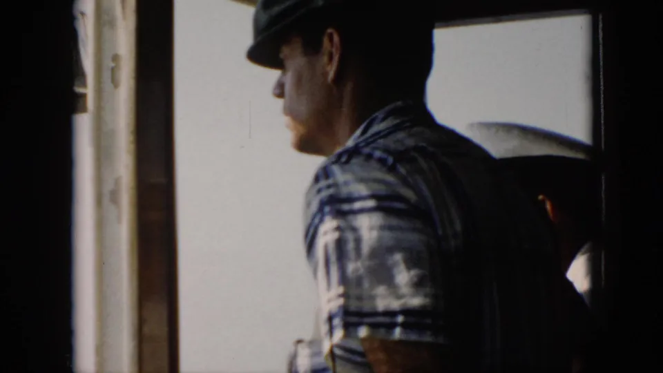 A passenger in a plaid shirt and cap sits in profile near a boat window during a 1956 Cuba water crossing.