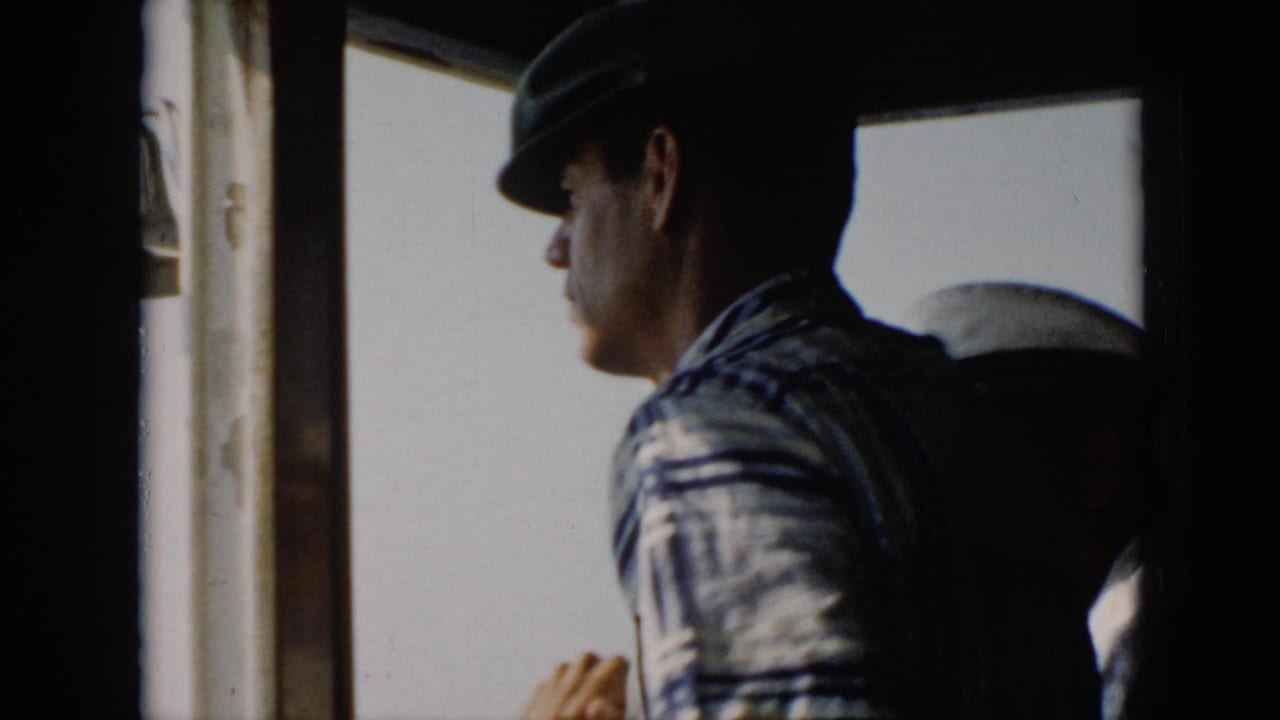 In 1956 Cuba home-movie footage, a man in a dark hat and striped shirt stands in profile inside a boat wheelhouse.