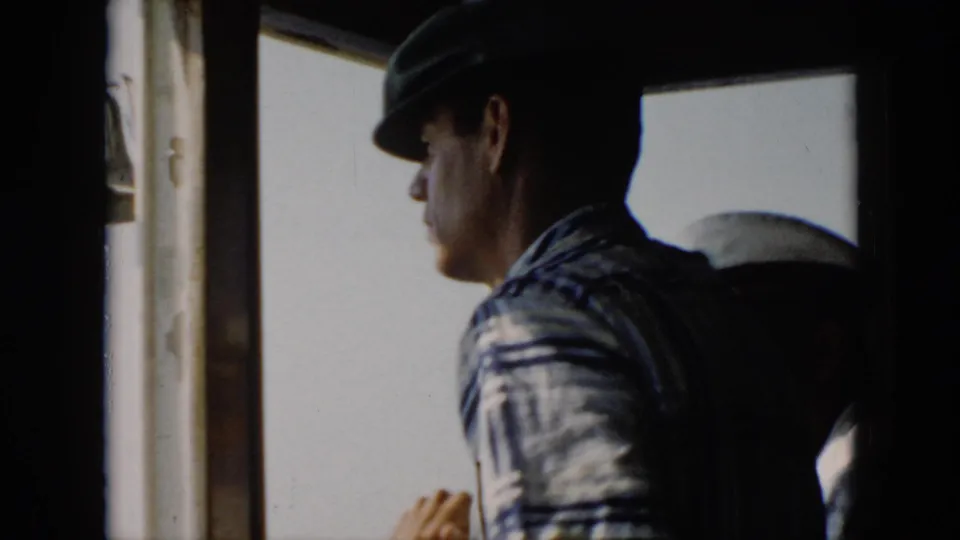 In 1956 Cuba home-movie footage, a man in a dark hat and striped shirt stands in profile inside a boat wheelhouse.