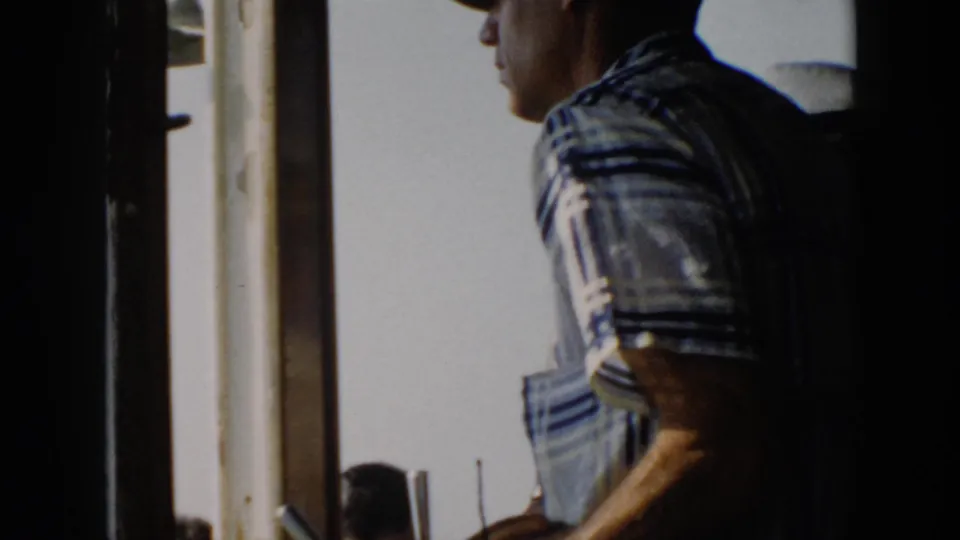 A man in a striped shirt stands by the steering wheel inside a boat, with bright daylight through the wheelhouse window.