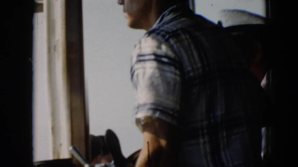 The camera catches the man from behind in the boat wheelhouse, with a second person in a white cap partly visible nearby.