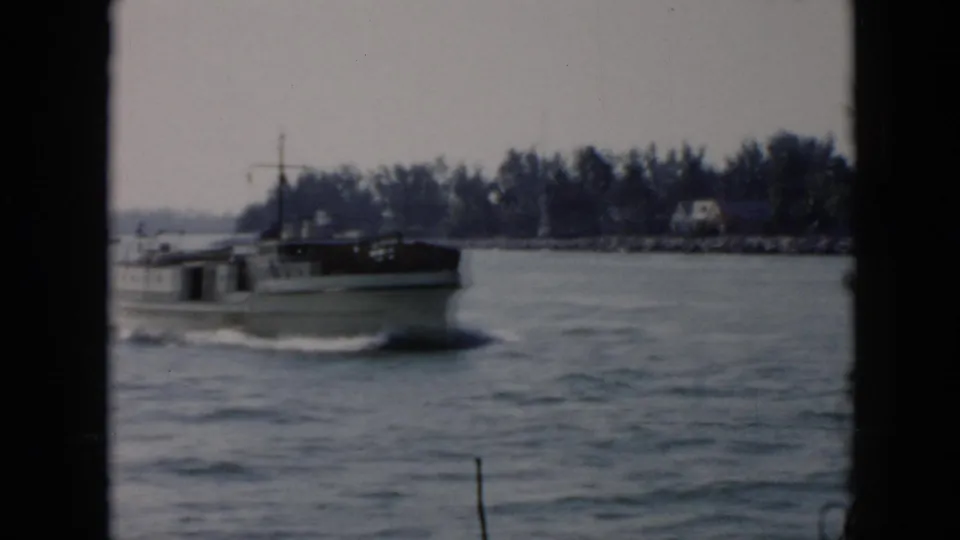 A white cabin motorboat passes near the camera on choppy water in Cuba, 1956, with trees and small shore buildings behind it.