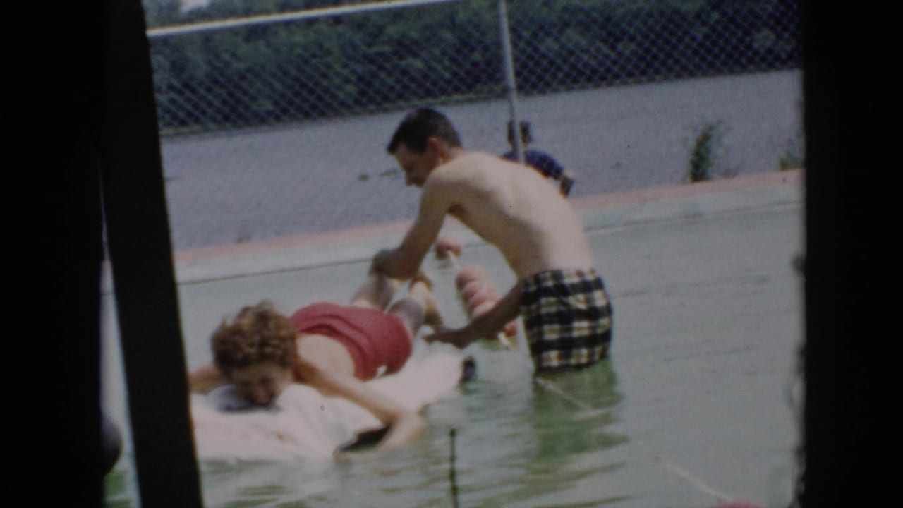 A young man in plaid trunks tickles another man lying on a pool raft.