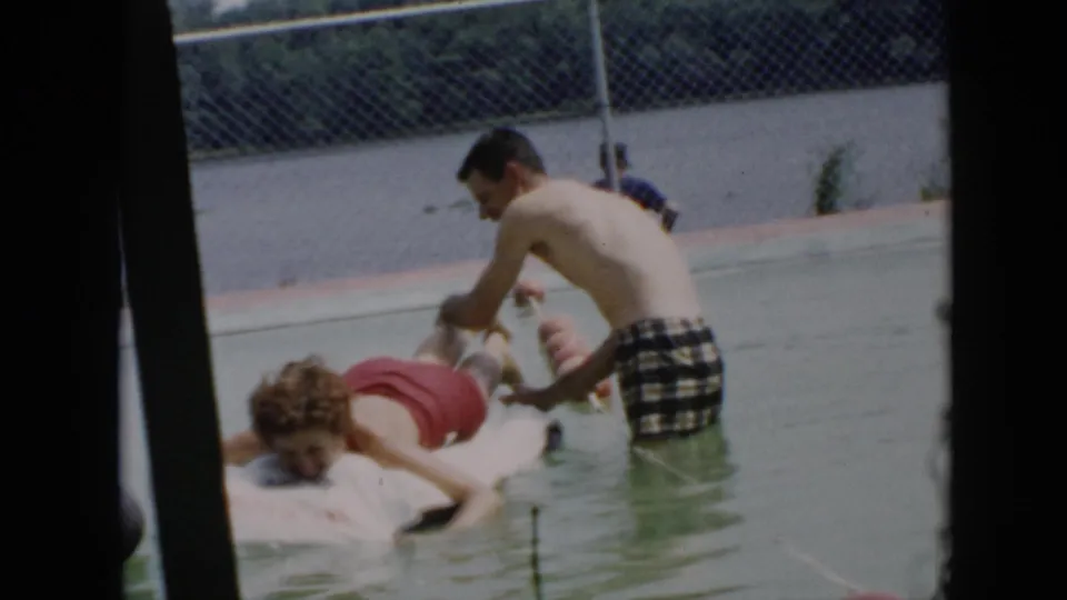 A young man in plaid swim trunks playfully tickles or pushes another man on a white raft in a Cuba swimming pool scene from 1956.
