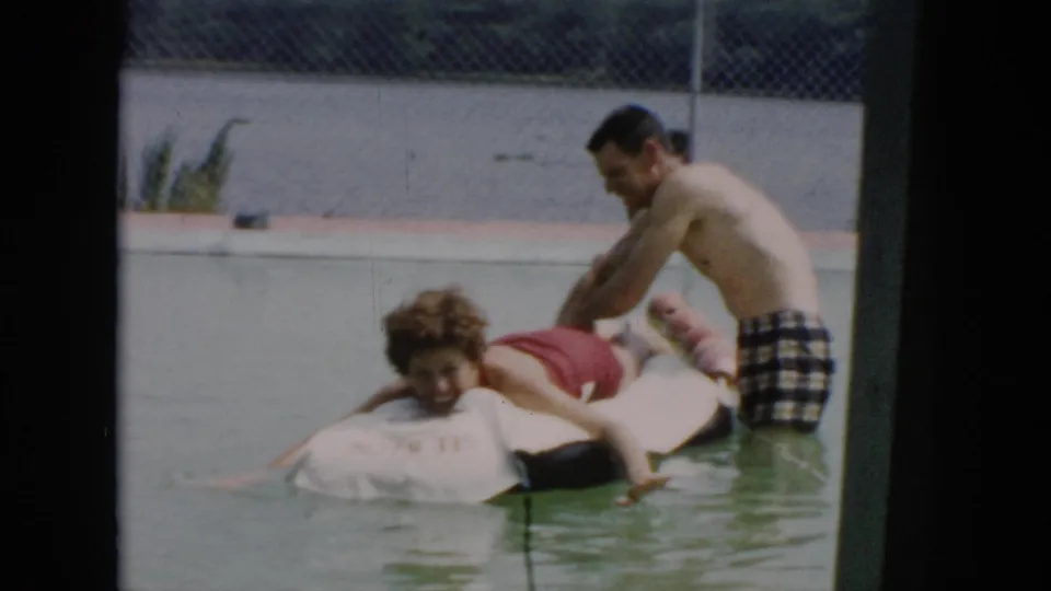 The man on the raft turns toward the camera as the standing man continues playful horseplay in the 1956 Cuba pool footage.
