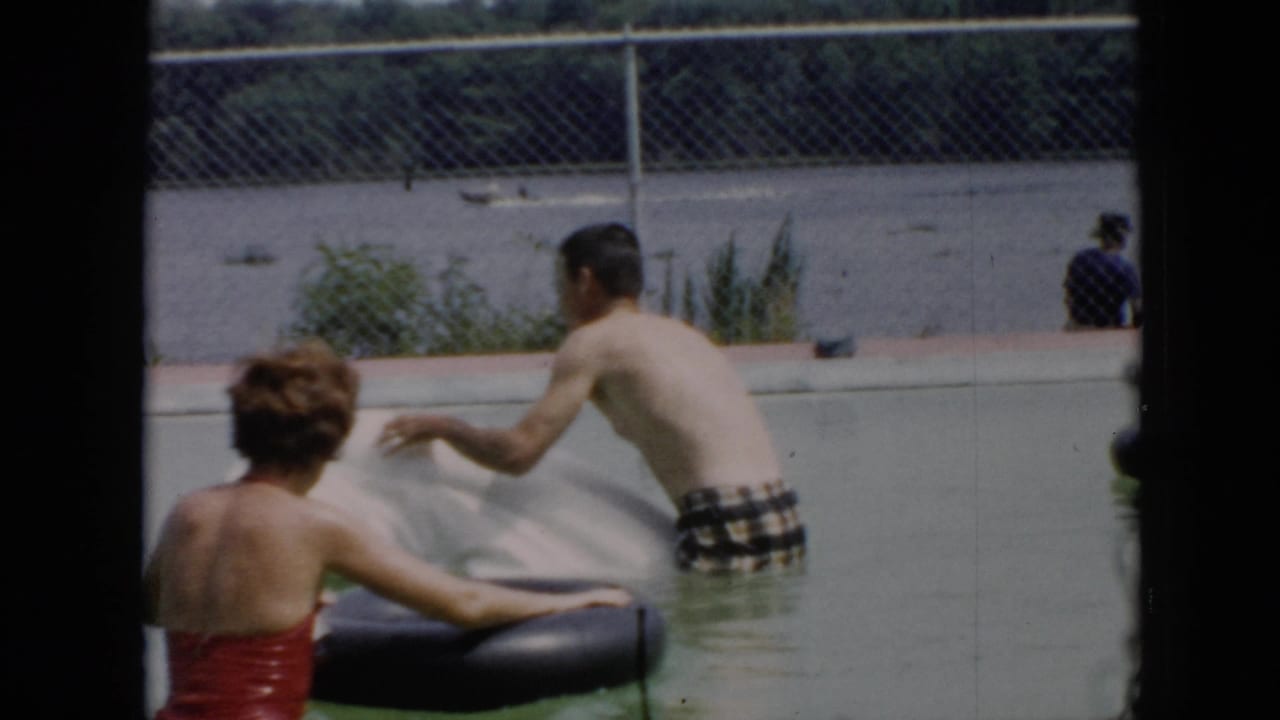 A man in plaid trunks reaches toward a white raft while a woman in a red swimsuit holds a black inner tube in the pool.
