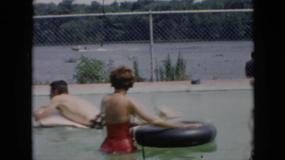 The man begins climbing or jumping onto the white inflatable raft as the woman in red remains near a black inner tube.