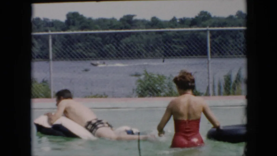 In the pool, the man lies stretched across the white inflatable raft while the woman in red turns near the black inner tube.