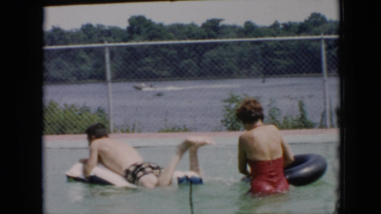 In a 1956 Cuba home-movie frame, a man floats face down on a white inflatable while a woman in a red one-piece swimsuit sits nearby with a black inner tube in an outdoor pool by a fenced waterway.