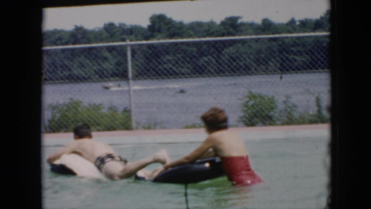 The 1956 Cuba pool scene shows the woman beside a black inner tube as she leans toward the man on a white inflatable, with a chain-link fence, water, and trees behind them.