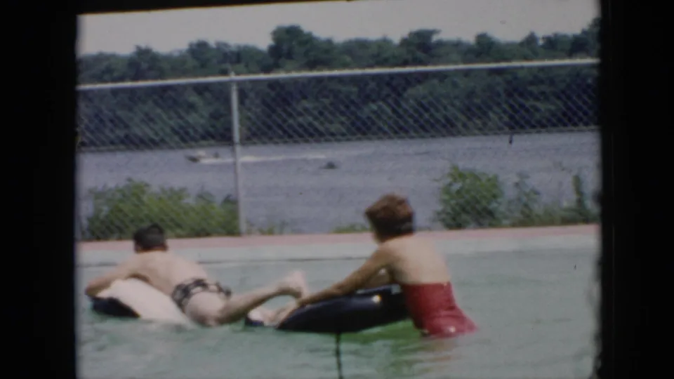 The 1956 Cuba pool scene shows the woman beside a black inner tube as she leans toward the man on a white inflatable, with a chain-link fence, water, and trees behind them.