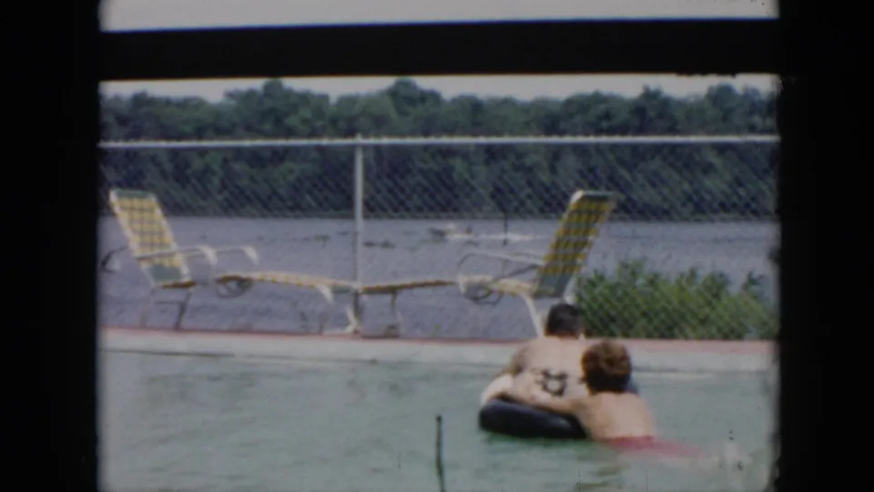 A 1956 Cuba home-movie frame places the couple lower in the pool beside a black inner tube, with yellow lounge chairs along the deck and a lake or waterway visible beyond the chain-link fence.