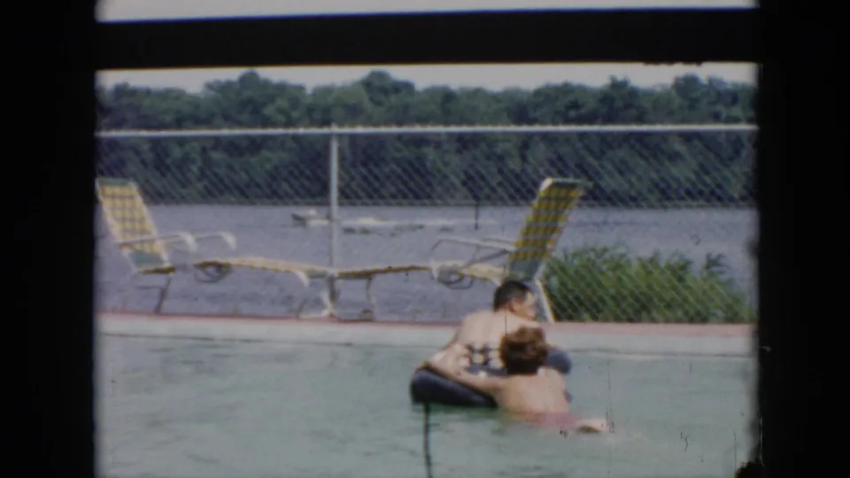 In the final 1956 Cuba frame, the couple remains in the pool around a black inner tube, backed by metal lounge chairs, a chain-link fence, and the broad waterway beyond.