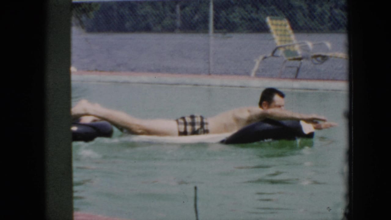 Man lies across a black inner tube in a pool with lounge chairs behind a fence.