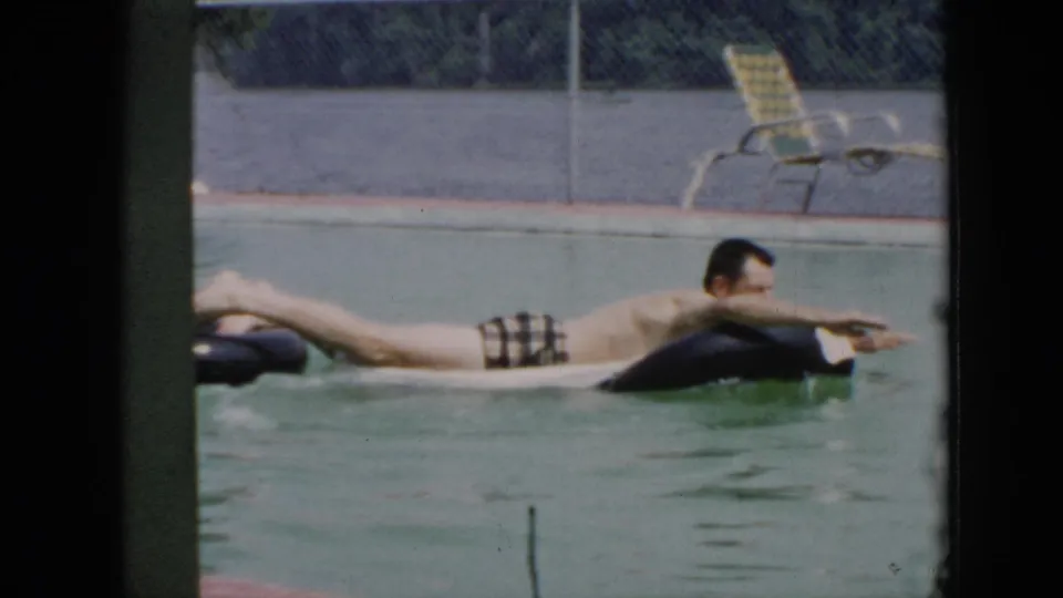 The man continues floating across a black inner tube in the pool, with a fence and lounge chairs in the background.