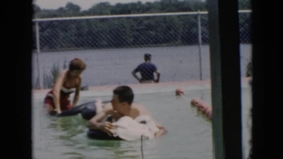 A woman in a red and white swimsuit stands in the pool beside men on inflatable floats during a casual family moment.
