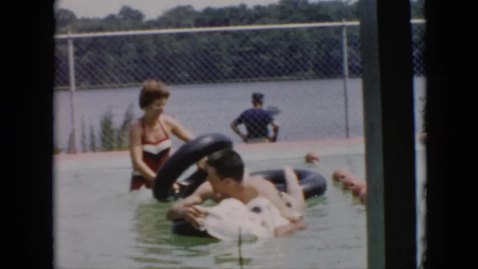 A woman holds a black inner tube near a reclining man in the pool, with another adult seated by the fence behind them.