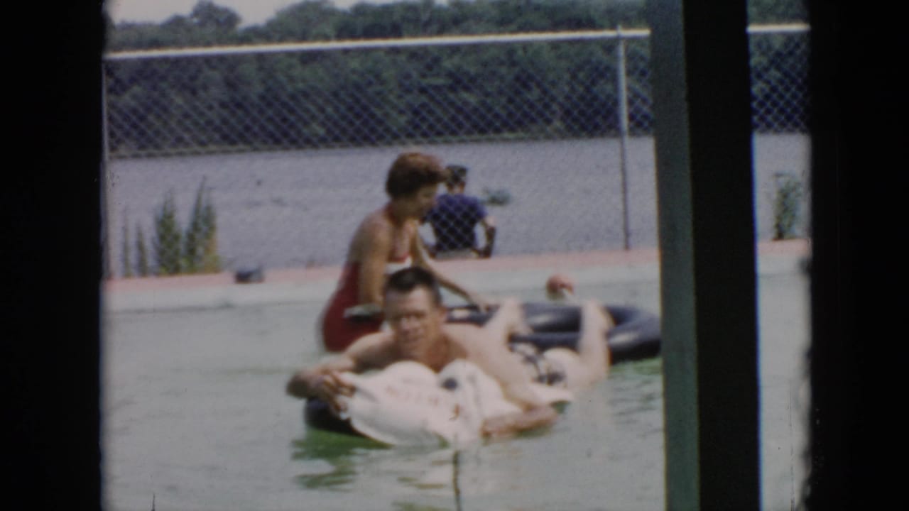 1956 Cuba home-movie frame showing adults on inflatable tubes near a chain-link fence, viewed through dark vertical framing.