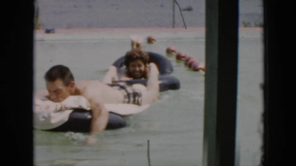 1956 Cuba frame of adults floating on black inner tubes in calm water, seen through a window or structural frame.