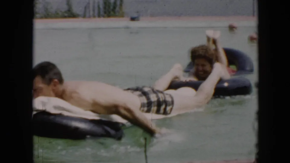 Close 1956 Cuba home-movie view of adults on inflatable tubes, with soft focus, lake water, and dark frame bars at the sides.