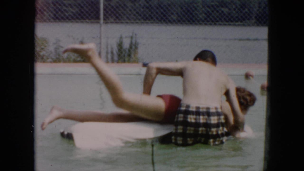 A person kicks a leg upward from a white float while another person supports them in the pool.
