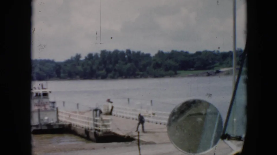 A 1956 Cuba ferry landing appears through a vehicle window, with a worker on the pier and the river behind.