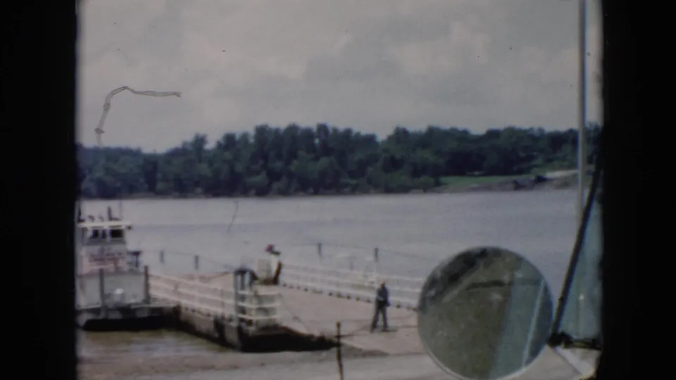 1956 Cuba ferry dock scene with workers near mooring lines, a vehicle mirror in the foreground, and trees across the river.